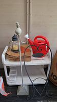 White plastic two-shelf garden cart with items on top shelf including glass candle lantern, brown leaf bags, black metal heart-shaped welcome sign, red watering can, and a tall white metal stand.