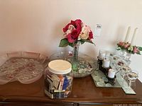 Photo of various decorative items including a large floral glass serving plate, faux flower arrangement in a glass vase, and several candles in glass holders on a mirrored tray.