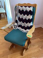 Wooden glider rocker chair showing angled armrests and green fabric cushions with crocheted afghan draped over