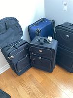 Four luggage pieces arranged in corner: two black soft-sided suitcases with wheels, one black carry-on bag without wheels, and one dark blue hard shell suitcase with wheels.