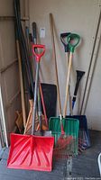 Photo showing multiple outdoor tools including snow shovels with red, green, and blue plastic scoops, metal and wooden rakes, a shovel and an edger.