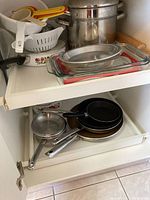 White kitchen cabinet shelf containing various fry pans, cooking pots, glass baking dishes, plastic measuring cups and a colander.