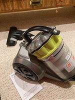 Hoover MultiFloor vacuum cleaner with transparent dust bin and black handle on beige carpet, includes instruction manual beneath the vacuum and black floor cleaning head attached.