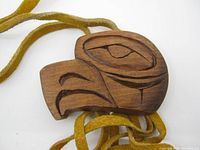 Close-up front view of hand carved wooden pendant showing Indigenous style design with eye and beak imagery, attached to leather cords.