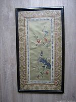 Full view of framed embroidery picture showing bird and flower design horizontally vertical in black frame on gray wood floor.