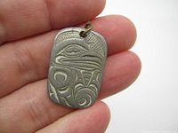 Pewter pendant held in fingers showing embossed raven design with indigenous art patterns.