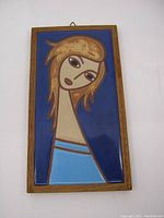 Frontal view of rectangular ceramic tile featuring stylized portrait of woman with gold hair and blue dress in wooden frame.
