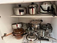 Wide view of cabinet shelf with various pots and pans including glass pots and stainless steel cookware plus Tatung rice cooker.