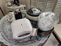Photo showing all five small kitchen appliances on a round table with lace cloth: Toastmaster sandwich maker, Black and Decker handy chopper, Hamilton Beach can opener, Black and Decker cooker, Tiger Hot Pot
