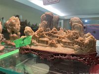 Front and side view of the soapstone carving showing mountainous landscape, traditional houses, trees, and the detailed wooden base.