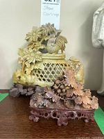Two hand carved soap stone sculptures; the smaller purple floral and grapevine carving is foregrounded with a larger yellowish rooster floral lattice carving in the background.