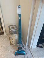 Full view of upright Electrolux cordless vacuum cleaner in blue color leaning against wall next to unopened household items.