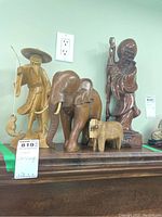 Four wooden carvings displayed together on a wooden surface: an elephant, fisherman, wise man, and hippo, under a light switch.