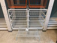 Two metal storage units with three wire baskets each shown side by side. One unit has orange wire baskets on top and white on bottom, the other has all white baskets. Units have metal frames and sit on tile floor.