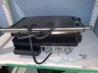 Full view of Breville indoor electric BBQ grill with stainless steel handle, power cord wrapped around handle, temperature control knobs, and drip tray.