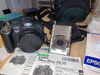 Front view showing both Canon cameras, manuals, photo paper, and carrying bag on table.