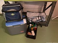 Camping gear items including Coleman cooler, Arctic Zone lunch bag, blow up mattress, mosquito repellents, clothes pins on a basement shelf and floor.
