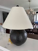 Front view of the table lamp showing the spherical black ceramic base with crackled pattern and large white conical shade, standing on a white surface with background of dining room furniture.