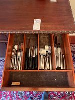 Photo of opened wooden drawer showing cutlery set arranged by type. Four compartments hold spoons, steak knives with black handles, forks, and regular knives.