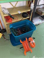 Bin filled with assorted extension cords, two orange extension cords coiled externally, and two work lights on metal shelving