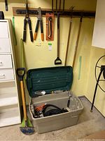 Garden tools hung on wall including shears, loppers, pruning saw, garden rake, long-handled tool, and large beige tote bin with lid on floor containing garden supplies and coiled hose.