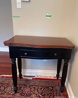 Front view of the folding table, showing brown top with scalloped edges, black base, one drawer with metal pulls, turned legs on a patterned rug.
