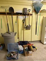 Wide shot showing garden tools mounted on a yellow wall including rakes, shovels, and snow shovel, next to a Rubbermaid garbage bin and storage box on floor.