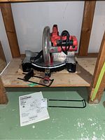 Red and black Craftsman compound miter saw on wooden shelf, including instruction manual and two metal stand legs on floor.