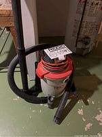 Front and side view showing the Ridgid WD0600 shop vacuum with hose, power cord wrapped on top, and owner's manual placed on top. The shop vac is positioned on the basement floor near a water heater.
