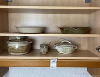 Two ceramic pie plates on a shelf, one plain beige and one with green glaze. Below are two lidded handled bowls and a small bowl with a spoon.