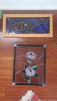 Two stained glass panels, one framed with wood featuring a fish design, the other square with flowers and hanging chain