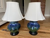 Pair of ceramic base table lamps with white fabric shades shown together on wooden floor. Black wooden base with blue-green glazed ceramic middle. Electrical cords with inline switches visible.