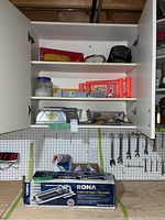 Photo of white cabinet open above the workbench showing painting supplies, a hat and goggles, and the tile cutter box beneath it.