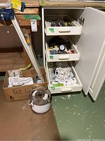 Photo showing the white cabinet with three drawers partially open, wire spool on floor, box labeled 'Pot L', and other hardware items around.