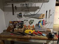 Wide shot of workbench holding various hand tools against pegboard wall. Shows handsaws, boxed drywall tools, clear plastic containers, and smaller hand tools.