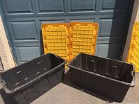 Two large black rectangular storage containers with yellow snap lids placed behind them, showing the interior pattern of the lids and handles on the containers.