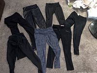 Seven pairs of women's dress pants on floor, six black and one blue, showing variety of styles and materials.