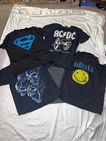 Five black men's large T-shirts on white backdrop: Superman logo, AC/DC band shirt, Nirvana smiley face, smoky skull with flames, and black/red graphic design.