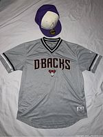 Gray Arizona Diamondbacks jersey with black and white sleeve stripes and front team logo laid flat on surface with purple World Series cap placed above jersey.