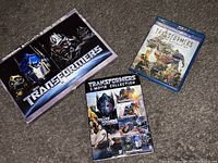 Photo showing three Transformer movie discs: a boxed 2-disc DVD set, a 5-movie DVD collection, and a Blu-ray disc of Transformers: The Last Knight.