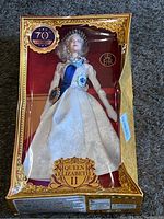 Full view of the boxed Queen Elizabeth II Barbie doll showing the white patterned gown, blue sash, tiara, and gold ornate box with 70th anniversary labeling.
