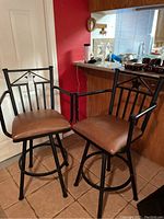 Pair of black metal swivel bar stools with brown vinyl padded seats shown side by side. Each stool has armrests, and the frames include a star design on backrests and circular footrests.