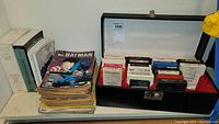 Overall view of box holding comics, books, and 8-tracks showing stacked Batman comics, books on shelf, and black case with 8-track tapes inside.