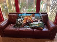 Front view of red-brown leather sofa with a lion print blanket and pillows on the cushions, positioned in front of glass windows.