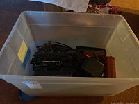 Plastic tote containing Lionel locomotives, train cars, curved metal train tracks, and control transformer photographed from above in natural indoor lighting.