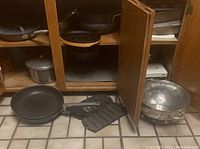 Cast iron frying pans including a griddle pan with ridges and a small cast iron pan behind cabinet doors next to metal colander on floor.