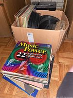 Photo depicting a large box filled with multiple 12-inch vinyl records and an assortment of album covers stacked outside the box, including 'Music Power' compilation