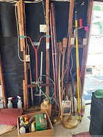 Full image showing the assembled yard and garden items leaning against garage wall, including tiki torches, pogo stick, sprayers, and umbrella stand.