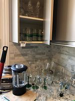 Image showing KitchenAid Architect blender jar and base on kitchen counter alongside multiple clear stemmed glasses and tumblers arranged around it, with some glasses inside an open cabinet.