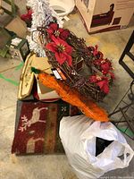 Photo showing grapevine wreath decorated with red poinsettias and pinecones, white bottle brush tree, orange mesh cone decoration, decorative Christmas mat, and bag with more items under it.